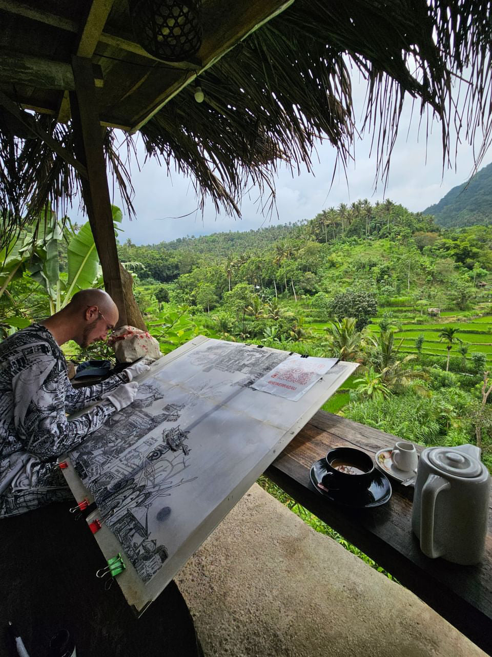 The artist is drawing a hand drawn illustration of Bali. He is sitting in a panoramic view with mountains and Reise fails. The artwork is black and white 